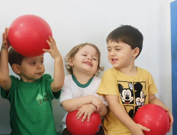 Três crianças sorridentes brincando com bolas vermelhas em um ambiente escolar acolhedor e descontraído, representando o clima alegre e inclusivo da Green Kids School, que combina imersão bilíngue com atividades lúdicas para incentivar aprendizado e autonomia.