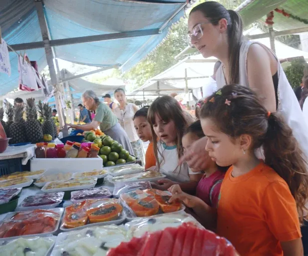 Educadora interagindo com crianças em uma feira ao ar livre, rodeada por frutas e legumes frescos, representando a abordagem integral e sustentável da Green Kids School em São Paulo, que une aprendizado, nutrição saudável e conexão com a natureza.