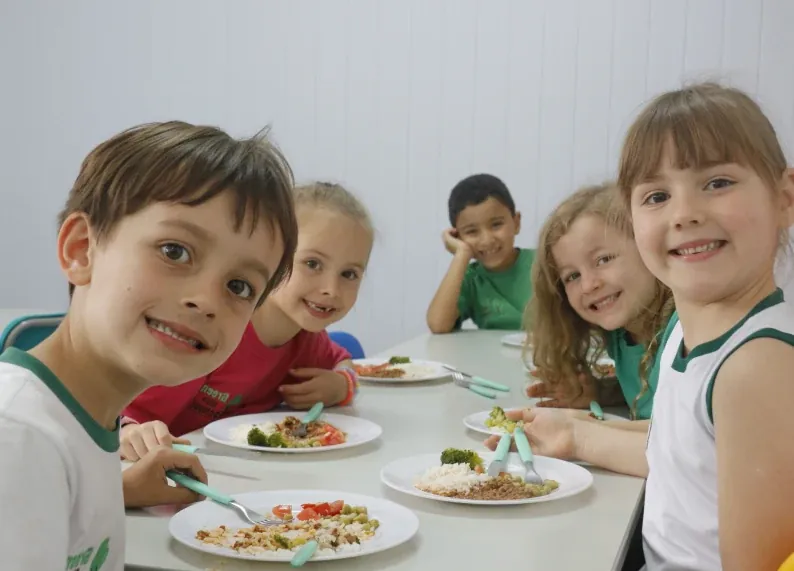 Um grupo de crianças sorridentes sentadas à mesa, degustando refeições nutritivas em um ambiente escolar acolhedor. A foto destaca o compromisso da Green Kids School com a educação nutricional e o incentivo a uma alimentação equilibrada.